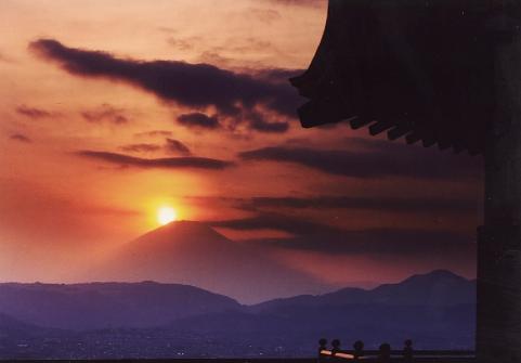 建物の軒先のシルエットの先に、山脈のシルエットがあり、その奥の富士山の山頂付近に沈む真っ赤な夕日が空を染め上げる幻想的なダイヤモンド富士の写真