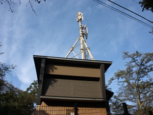 青空を背景に、木々の間に立つ通信機器用の建屋の屋根から、白い支柱で支えられた多数の無線アンテナが伸びる写真