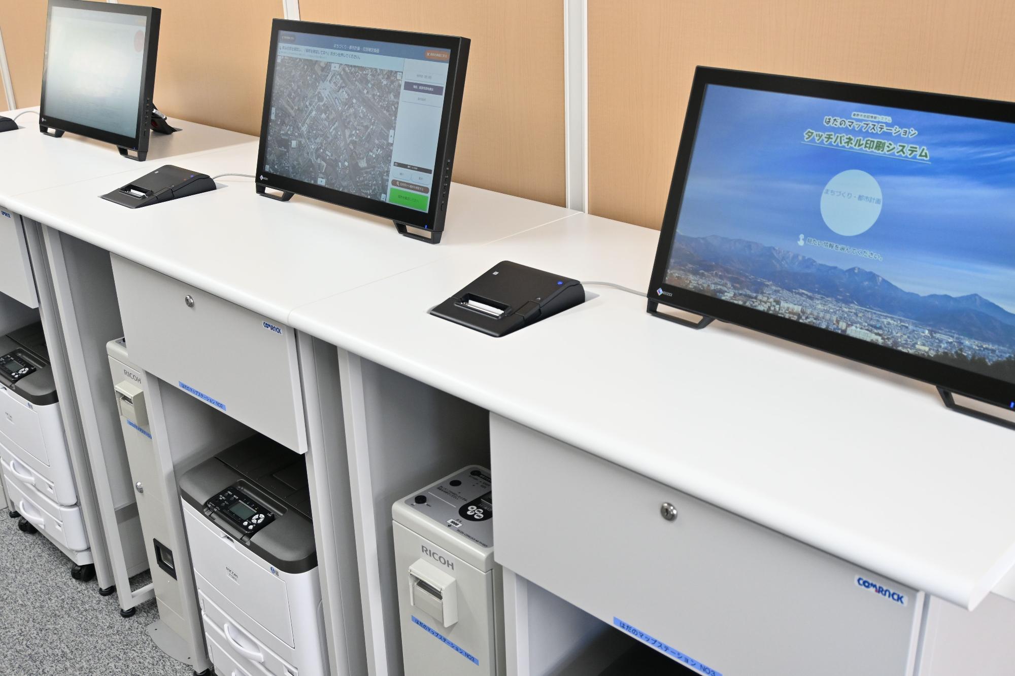 白いカウンターに複数の液晶モニターが並び、その下にプリンターが収納されたセルフサービス型の情報端末が設置されており、手前のモニター画面にはタッチパネル型証明書交付システムと表示されたマップステーションの写真