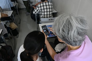 スマートフォン教室で白髪の女性が女子大生からスマートフォンの操作方法を教わっている様子を上から撮影した写真