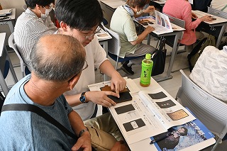 資料を机に置いた席に座った青いシャツを着た男性が、大学生の男性からスマートフォンの操作方法を教わっている、スマートフォン教室の様子の写真