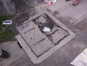 正方形のコンクリート製マンホール蓋が半分に割れ、中央部が大きく欠損し、内部の土やゴミが露出した側溝のふた破損の写真