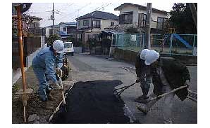 道路工事のヘルメットを着用した作業員たちが舗装された道路の一部に新しいアスファルトを敷き均している様子の写真