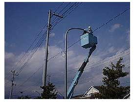 作業用の青い高所作業車がアームを伸ばし、バケットに人が乗って街灯のランプ部分を交換をしている様子の写真