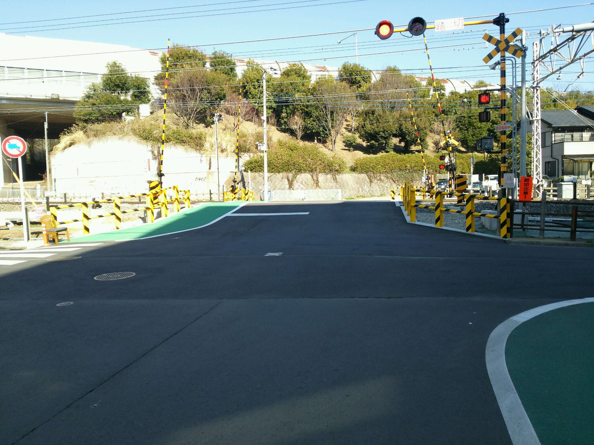 警報機が赤く点灯し遮断機が下りており、歩行者用道路が緑色に舗装され、見通しの良い踏切へ改良された全景を交差点から見た写真