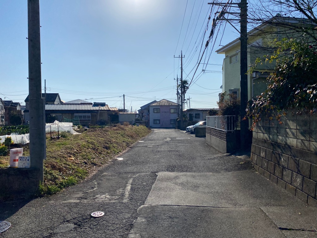 両脇には畑や電柱、平屋建てや2階建ての家々が立ち並んでいる工事前の市道80号線の写真