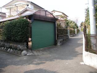 緑色のシャッターを備えたカーポート付きのガレージと、生け垣や石積みの基礎が見える住宅街の細い私道を撮影した整備前の写真