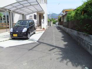 生垣などが取っ払われ透明な屋根がついたカーポートの下に黒い軽自動車が停まり、その横に二階建ての住宅が建つ、舗装された道がある整備後の写真