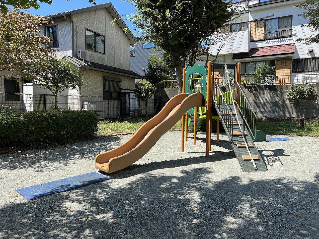 緑に囲まれた住宅地の中にある、明るい日差しの下に砂利が敷き詰められた小さな公園で、ベージュと緑色の複合遊具の滑り台が中央に配置されている様子の写真