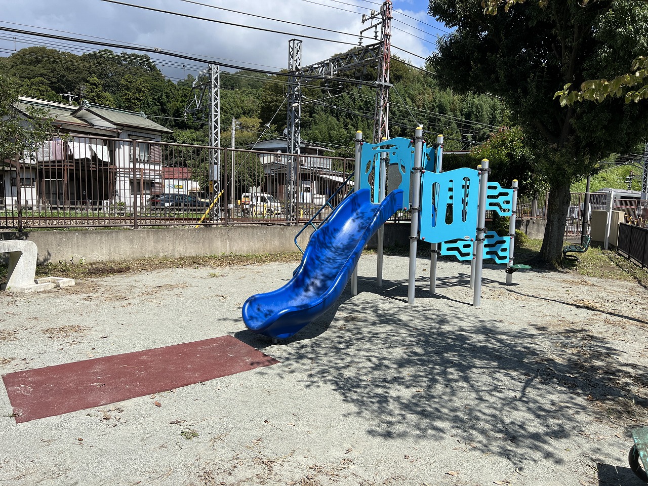 鉄道線路沿いの小さな公園に、青い滑り台とクライミングウォールが付いた水色の複合遊具があり、地面には砂利と茶色の衝撃吸収マットが敷かれている様子の写真