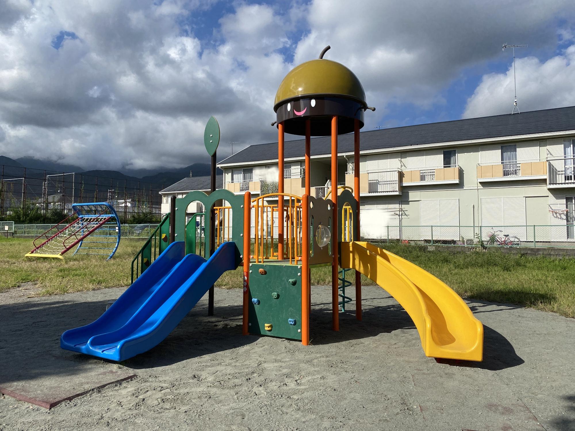 住宅や遠くの山々を背景に、砂が敷かれた広場の中央に、青と黄色の滑り台とクライミングウォールが付いたドングリの帽子をモチーフにした茶色のドーム型屋根を持つ遊具が配置されている写真