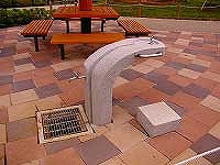 ブロック敷きの広場に設置された、木製のテーブルとベンチが設置された東屋の手前に、モダンなデザインの二つの蛇口が付いた石造りの水飲み場の写真