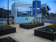 茶色い木製の花壇に色とりどりの花が植えられ、建物の前の低い壁には子供たちが遊ぶ様子が描かれたカラフルなイラストがあるくずわぶち美花（びか）公園の写真