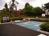 左側に公園の全体図と思われる看板、右側に通路がが設置され、手前に青色で駐車スペースが設けられた身障者用駐車場の写真