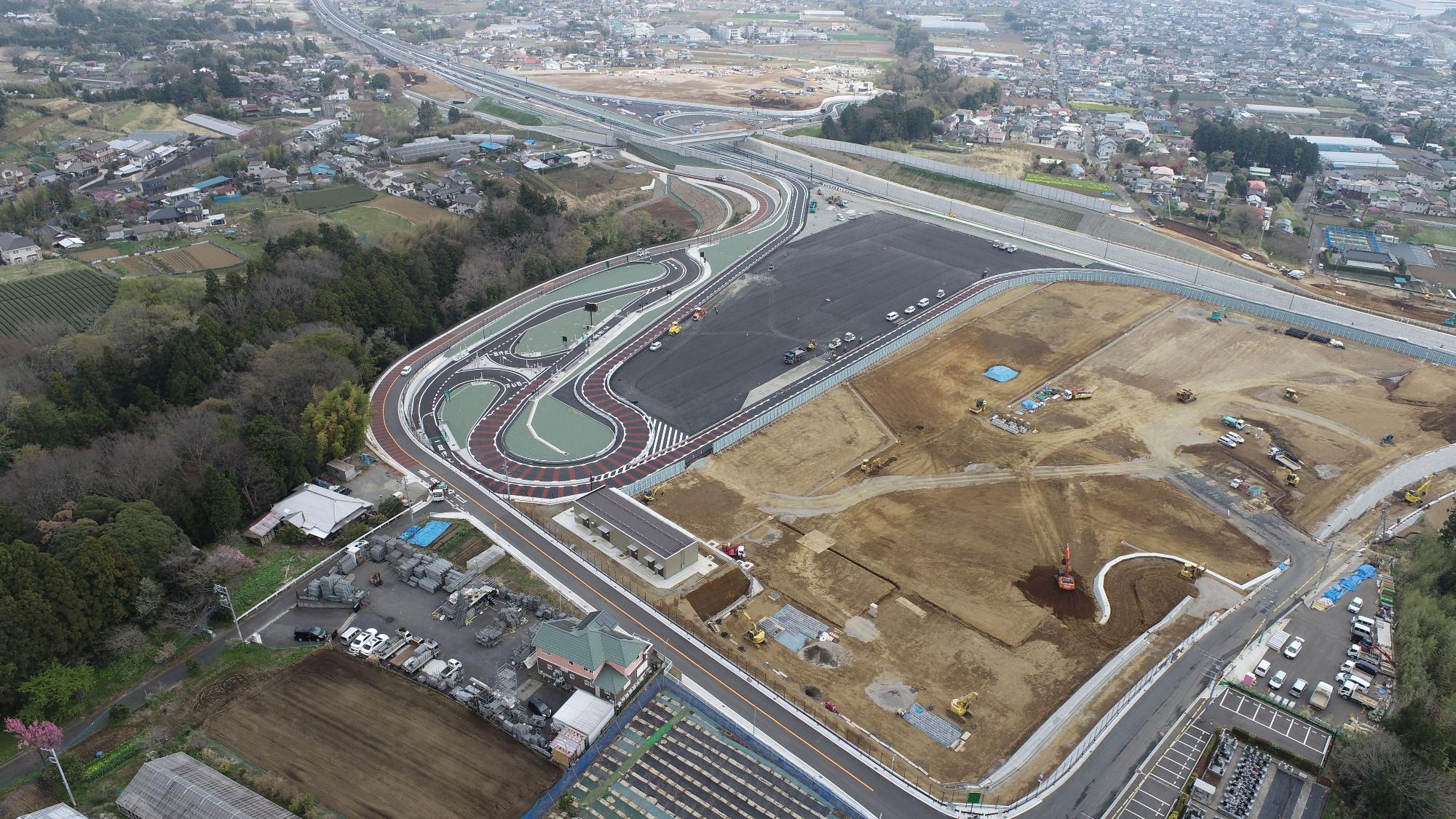 広大な造成地の建設現場と森の間に、緑色に色付けされたエリアがあり、その中をカーブが連続する茶色の道路が通り、奥の高速道路へ続いている、スマートインターチェンジ開通時の航空写真