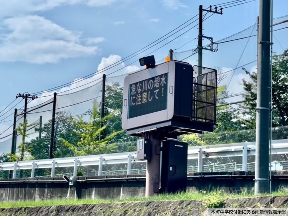 学校のネットを背景に、白いガードレールの外側の草地に設置された、「急な川の増水に注意して！」と表示がされた本町中学校付近にある雨量情報表示盤の写真