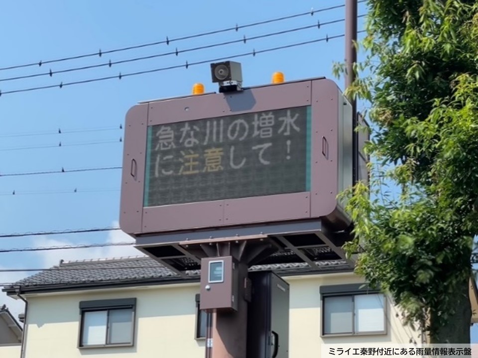 白い壁の建物の前に設置された、「急な川の増水に注意して！」と表示がされたミライエ秦野付近にある雨量情報表示盤の写真