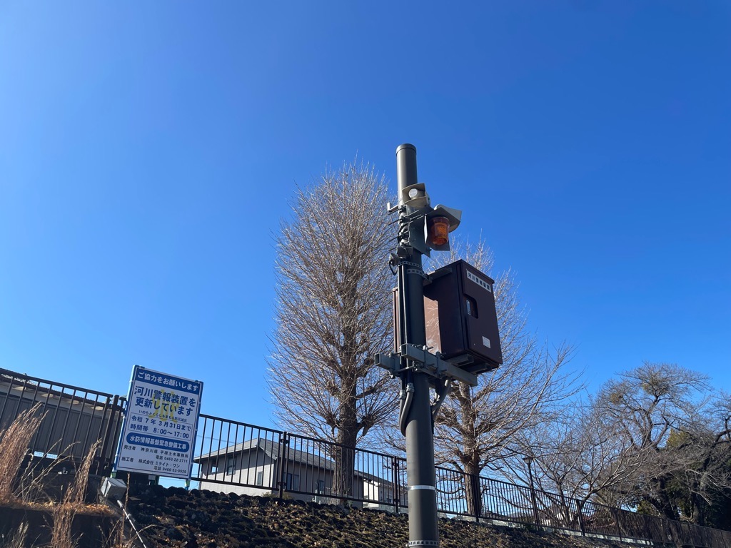 建物や木々の前にあるフェンスの下に法面が見え、その手前に、下親水施設警報装置が取り付けられたグレーの柱が立っている写真