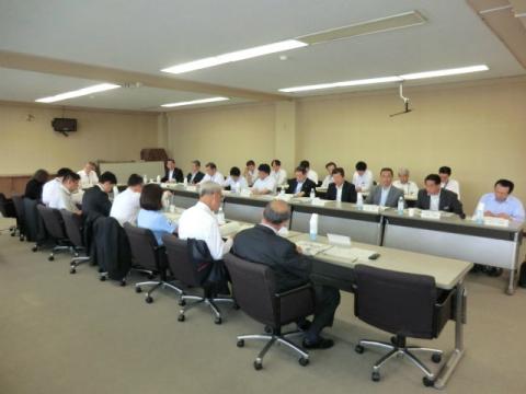 長い会議机をコの字型に配置した大会議室で、スーツやワイシャツ姿の数十人の出席者が着席し、真剣な面持ちで参加している会議風景の写真