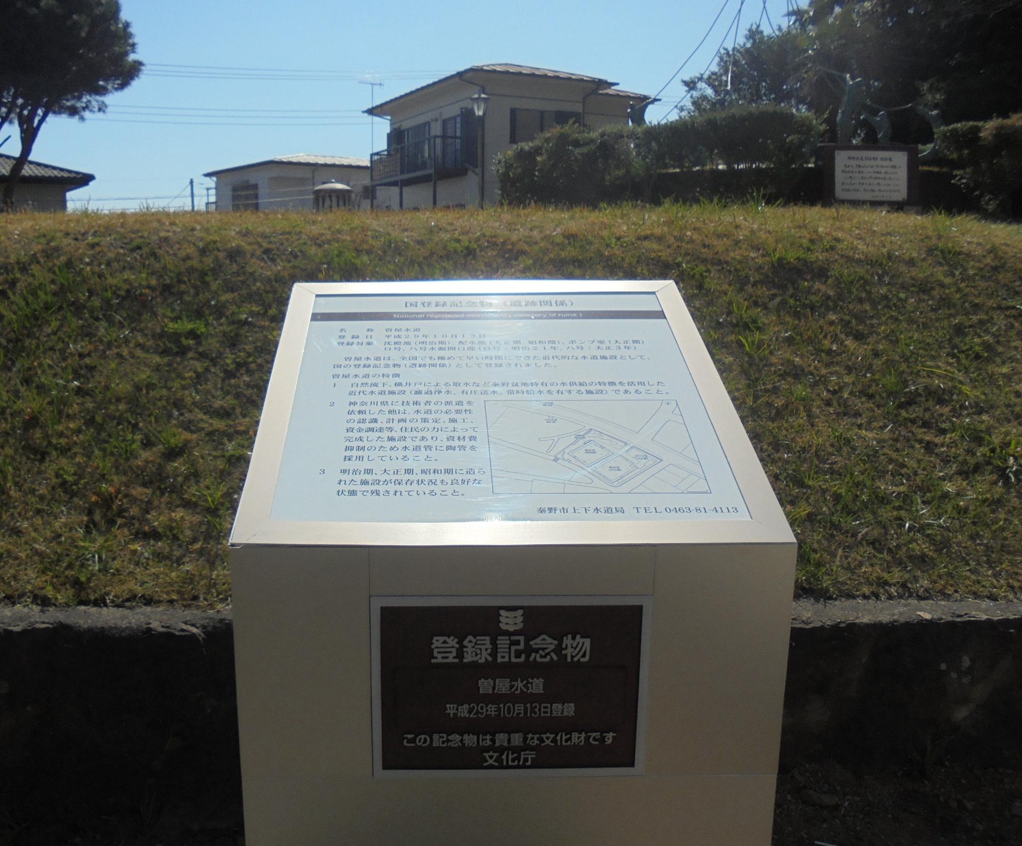 公園の芝生の法面の前に設置された白い四角の解説板に、曽屋水道の歴史が書かれている登録記念物の写真