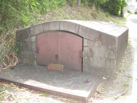 木々が密集した斜面下に設置された、セメントやコンクリートで出来たかまくらのような形をした小さな建物で、中央には茶色い金属製の扉がある現在のロ号水源の写真