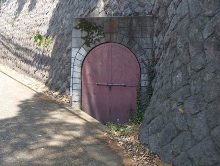 坂道沿いの石積みの擁壁に設けられた、茶色い金属製の扉が付いた、現在のハ号水源の入り口を捉えた写真