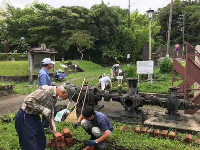 ボランティアの男性たちが、草が茂る公園の中で、古い機械の周りにレンガを並べたり、熊手で草をかき集めたりと、協力して環境整備をしている様子の写真