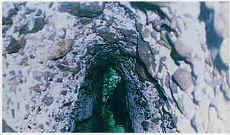 白くてゴツゴツした岩に囲まれた細い縦穴の隙間に、エメラルドグリーン色の水が深く溜まっている水源の写真