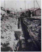 周囲に電柱が立ち民家が並んでいる中央に、深い溝が掘られている工事の様子の白黒写真