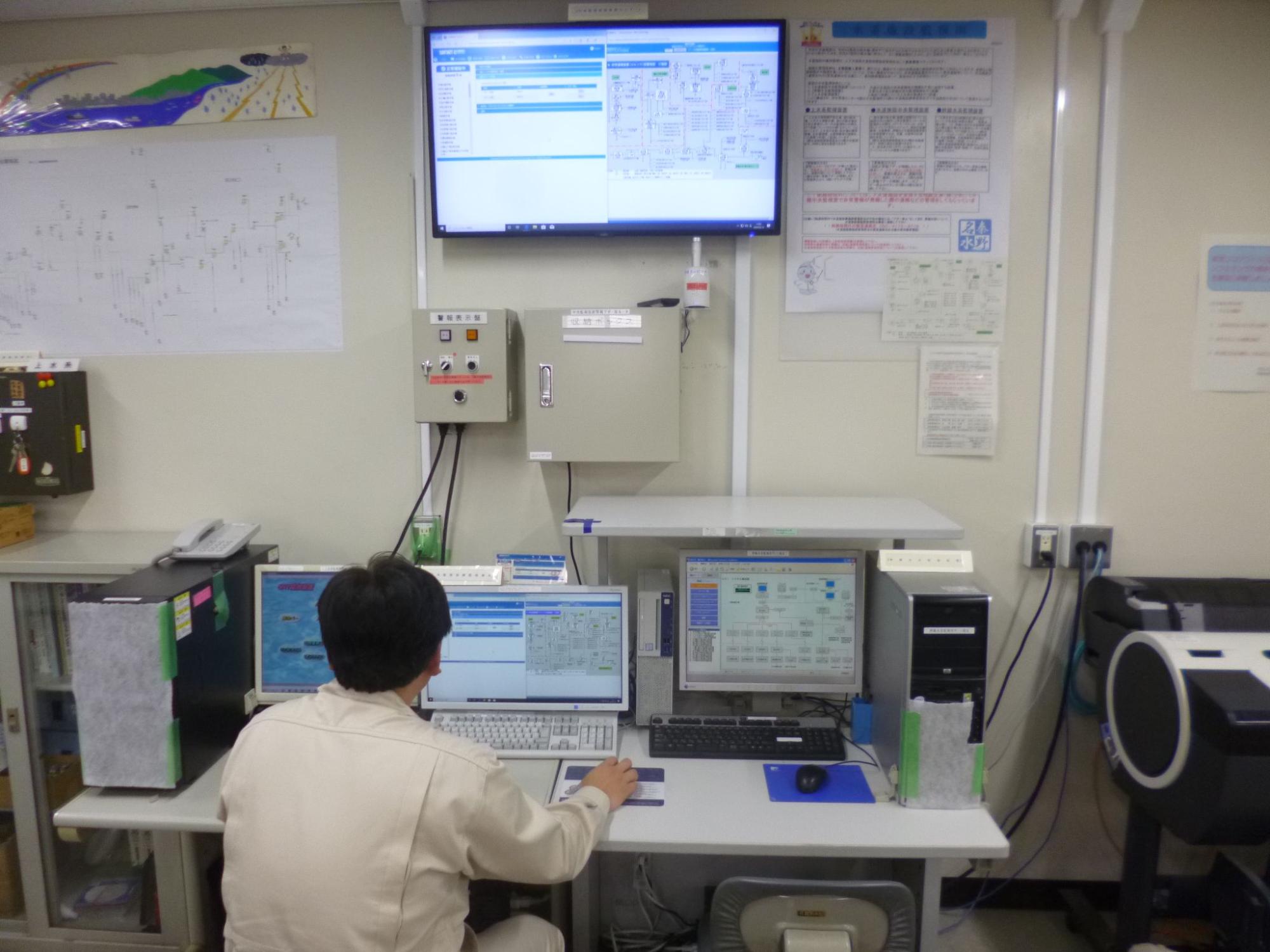 壁の大きなモニターとデスクにある複数のパソコンの画面にプラントの制御図やデータが表示されている監視室で、作業服を着た一人の職員がデスクに向かい、キーボードを操作している様子を後方から撮影した写真