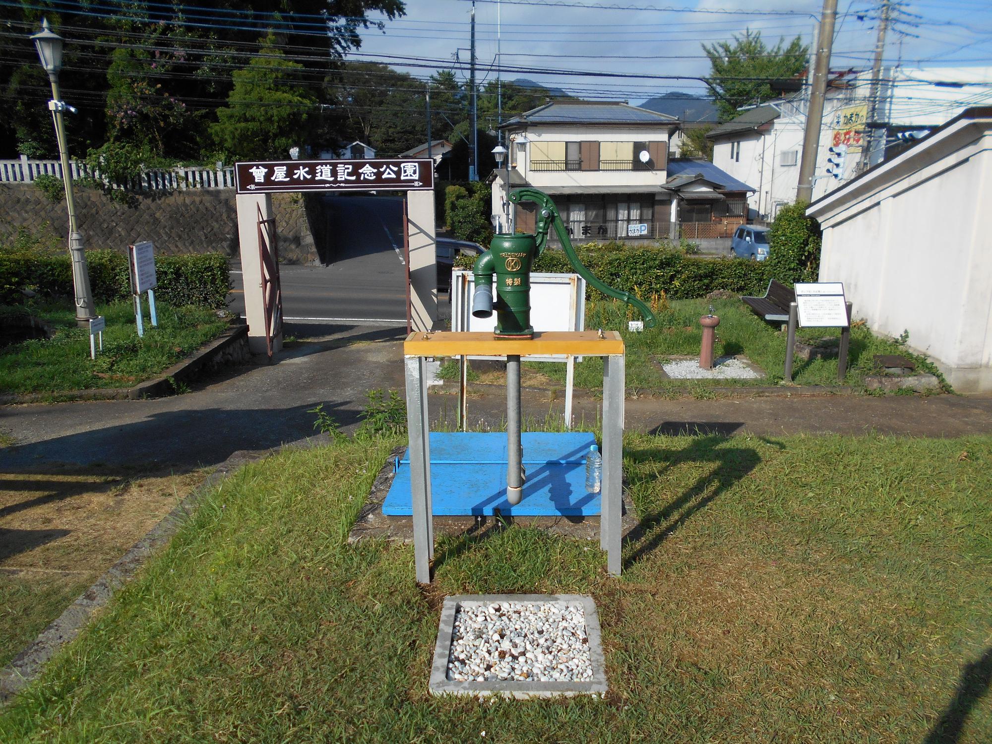 曽屋水道記念公園と書かれた入り口の門の前に、水色の鉄板が敷かれ、鉄製の台に載せられた緑色の手押しポンプが設置されている写真
