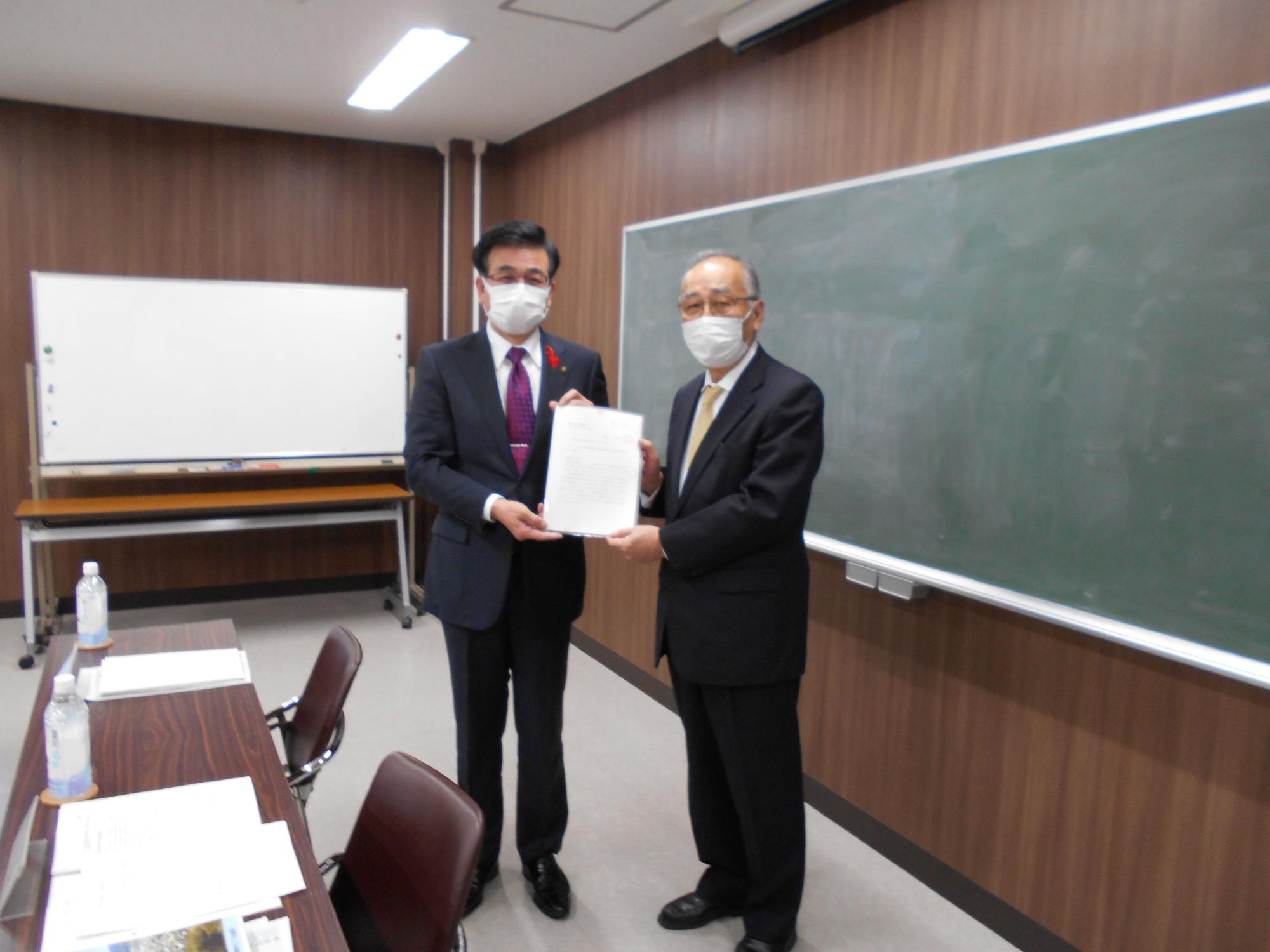 スーツを着てマスクを着用した市長と男性が、ホワイトボードと黒板のある会議室で、中央で1枚の紙を一緒に持ち、正面に向かって立っている写真