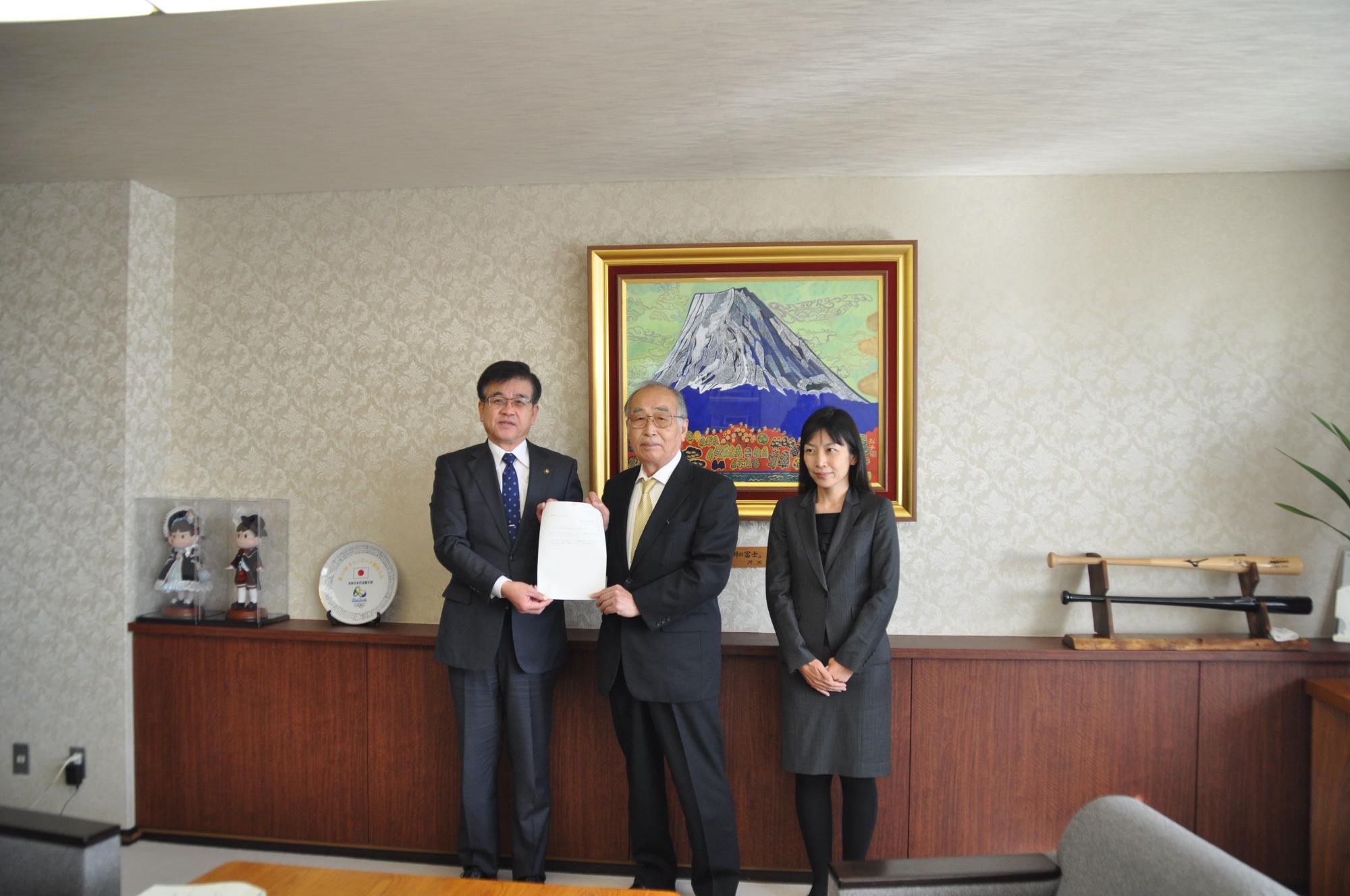 山の絵画が飾られた壁を背に並んで立った市長と、茂庭会長、松原副会長が答申書を持ってカメラに向かって立っている写真