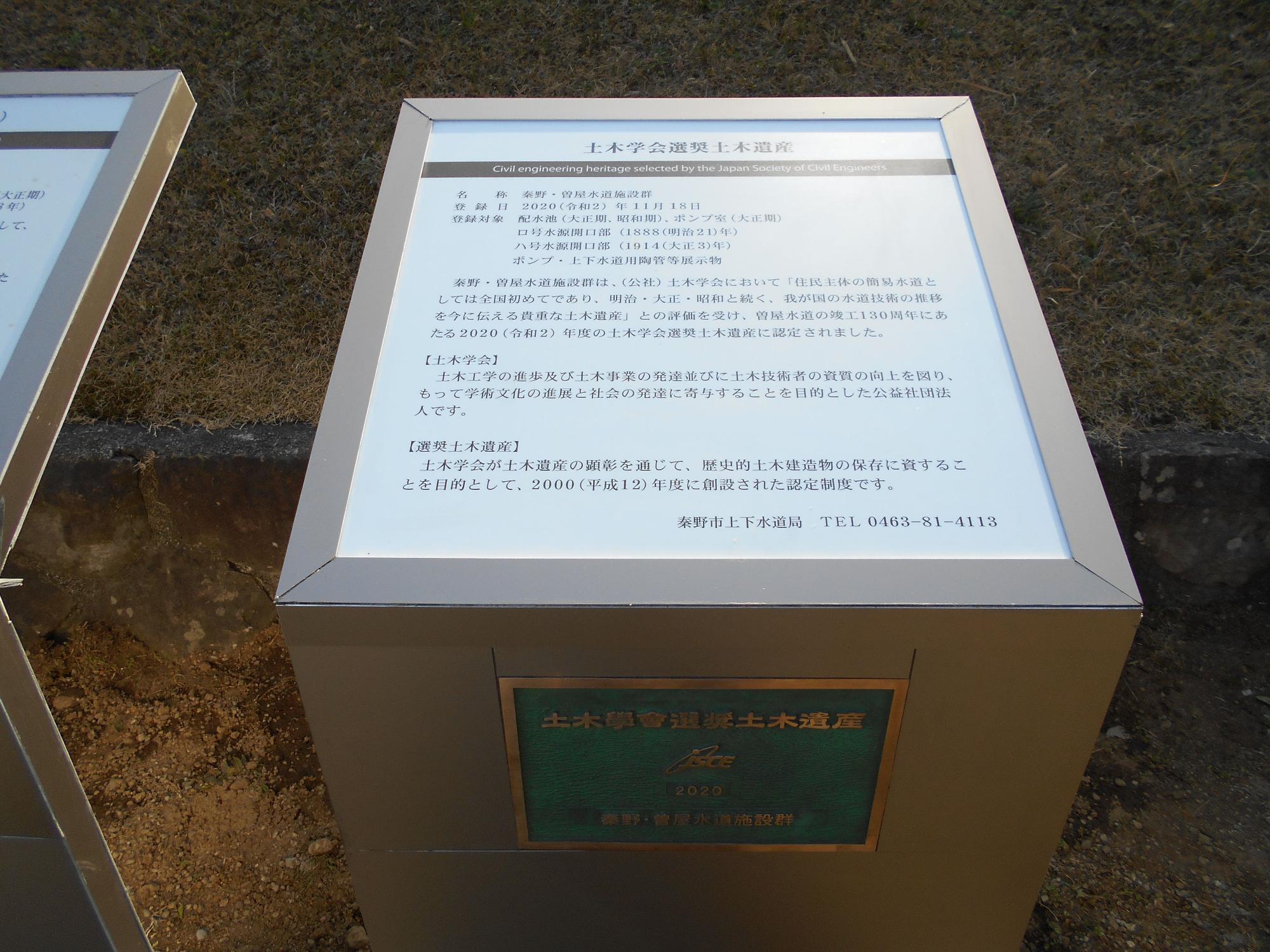 地面に設置された四角い案内板が写っており、上には説明文が記され、側面には緑色のプレートが設置されている土木学会選奨土木遺産の認定銘板の写真