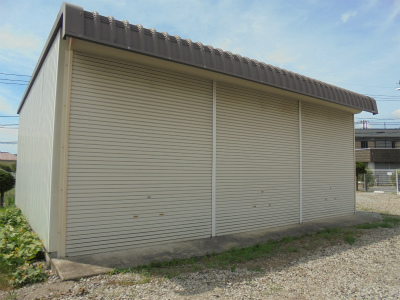 アイボリー色の金属製シャッターが横に三つ並んだ、波形の茶色い屋根を持つ車庫を正面からやや左側からの角度で撮影した写真