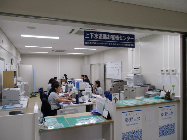 照明の明るいオフィスの一室で、手前にカウンター、その奥に複数のデスクがあり、パソコンに向かって座っている数名の職員が電話応対や事務作業を行っている様子を、入口付近から捉えた写真