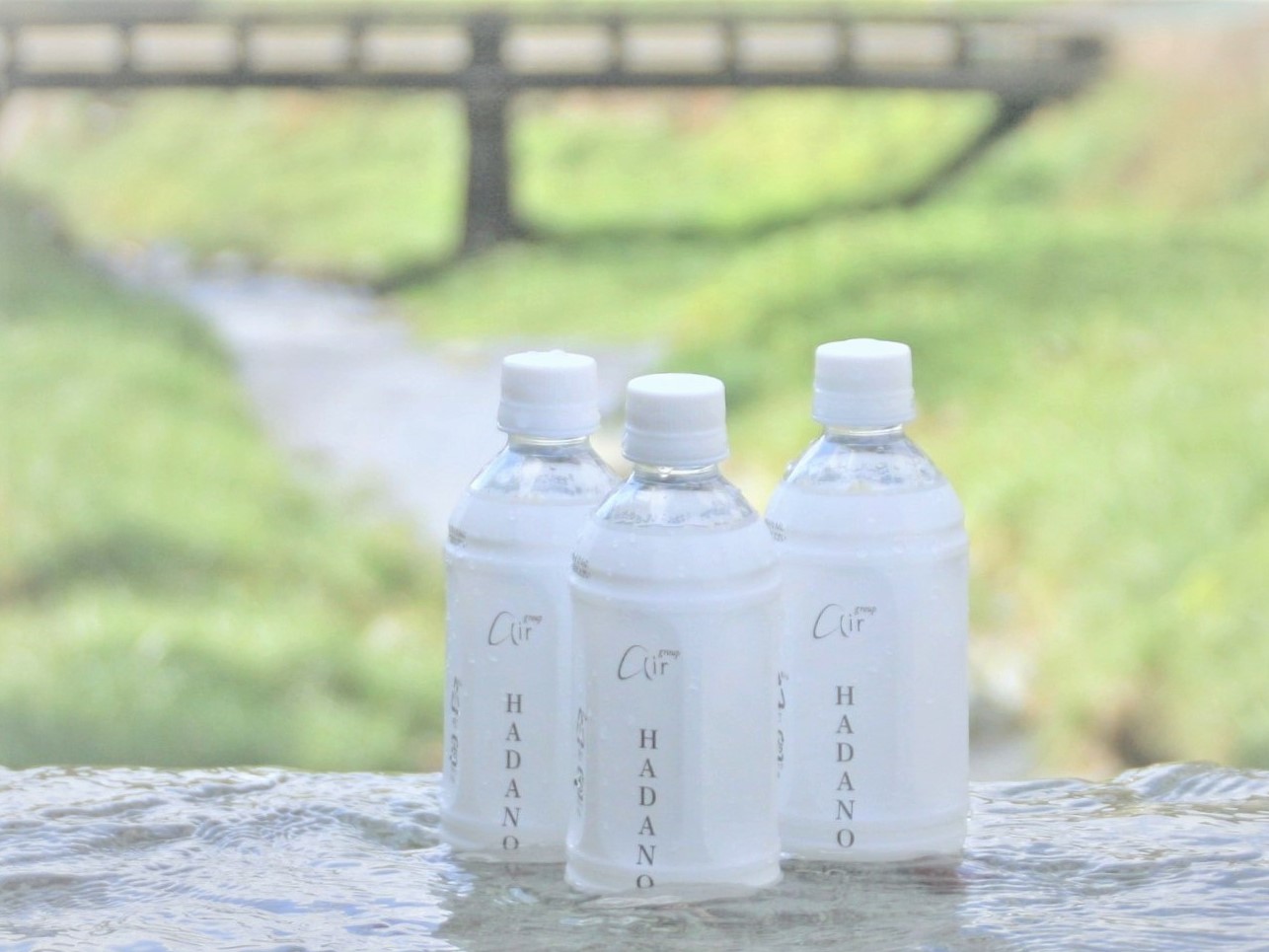 清流の手前に濡れた石のような場所があり、秦野のロゴと「Hadano」の文字が入った水のペットボトルが3本並んで置かれている爽やかなイメージ写真