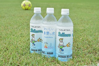 サッカーボールが背景にぼけている芝生の上に、湘南ベルマーレのマスコットキャラクターと秦野市のロゴが入った「おいしい秦野の水」のペットボトル3本が並んでいる写真