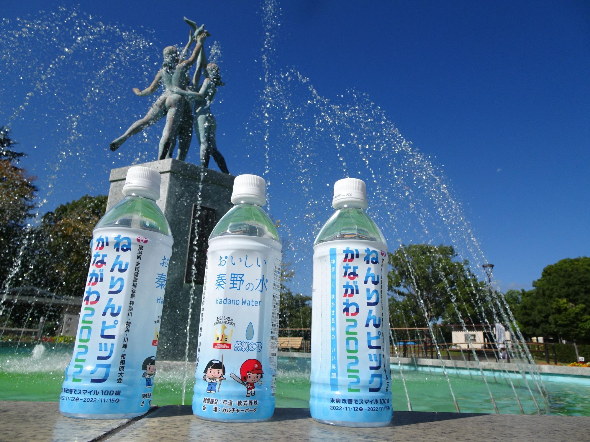 鮮やかな青空のもと、公園の噴水の前に、秦野市のマスコットキャラクター丹沢はだの3兄弟が描かれた「おいしい秦野の水」のペットボトル3本が並べられている写真