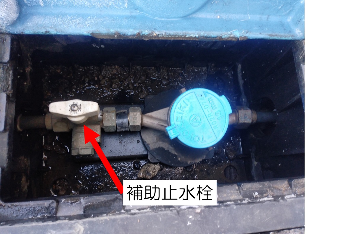 水道メーターボックスの内部が写されており、水色の蓋の付いた水道メーターと、赤い矢印で「補助止水栓」と示されたバルブが配管に設置されている写真