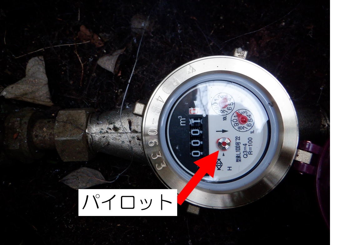 赤い矢印で「パイロット」と示された銀色のパイロットマークが写っている、薄暗い場所にある水道メーターをアップで撮影した写真