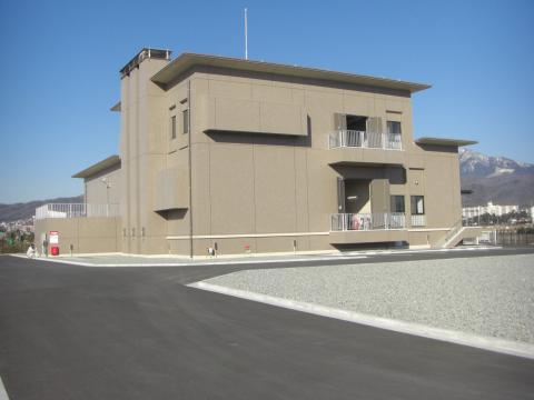 日当たりの良い青空の下、広々としたアスファルトの道路と砂利のスペースに囲まれた、平らな屋根と薄茶色の壁を持つ、無機質で近代的な設計の二階建てのポンプ場または制御施設の建屋を捉えた外観写真