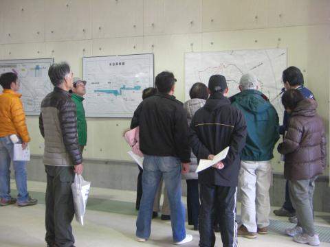 コンクリートの壁に貼られた複数の水路図や配管図の前で、厚手の防寒着を着た十数人の男女が、説明を聞いて見学している様子の屋内写真