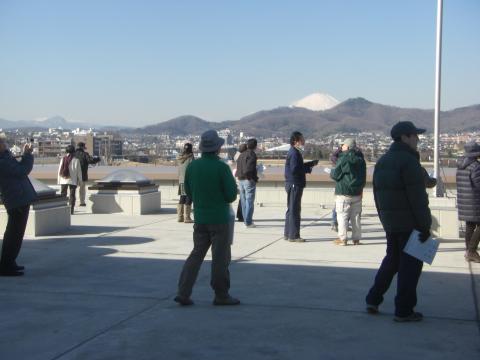 施設の屋上のような広いスペースに、防寒着を着た人々が数人集まっており、遠景には雪化粧をした富士山を含む山並みと街並みが広がる様子を捉えた見学風景の写真