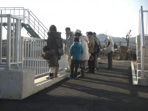 白い手すりと階段に囲まれた屋外の施設内で、防寒着を着た大人と子供のグループが説明を受けているように集まっており、遠くに街並みと山並みが見える見学風景の写真