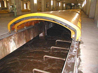 屋内に設置された長い水路に茶色い水が流れ、その上を黄色いアーチ状のカバーが覆っている写真