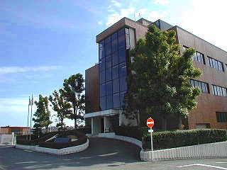 濃い茶色のタイル張りの外壁と大きな窓ガラスを持つ近代的な三階建て程度のオフィスビルを、その正面玄関への緩やかなカーブを描くスロープと整然と配置された植木や樹木、そして「進入禁止」の標識と共に捉えた写真