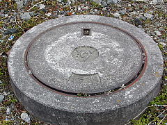 コンクリート製で上部に「汚水」の文字とマークが刻印された円形のマンホール蓋が、小石や雑草が生えた地面に設置されている様子を俯瞰で撮影した写真