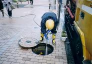 ベージュのレンガ敷きの歩道で、黄色のヘルメットと長靴、紺色の作業服を着た作業員が、ふたを開けたマンホールの穴にホースの水を勢いよく流し、清掃を行っている作業風景の写真