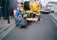 アスファルトの道路に設置されたマンホールの前に黄色の清掃車両が停車しており、その後ろで青い作業服を着た作業員2人が、車両後部から黒いコードを伸ばし、マンホール内の確認をしている様子の写真